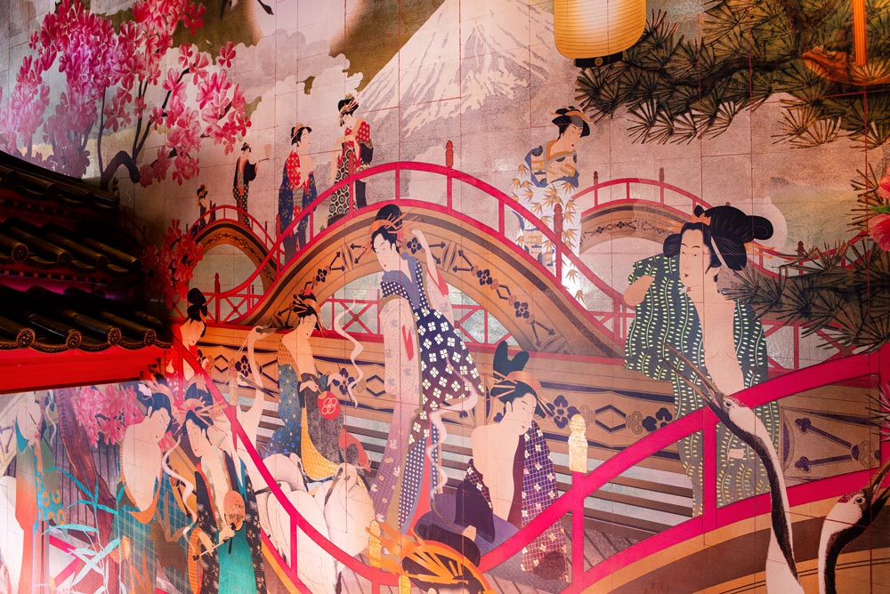 View across the entrance lobby bespoke wallpaper. Curved bridges with pink railings sit in front of a background of Mount Fiji. Pink cherry blossom trees are hand-painted and printed over the gold leaf metallic material. Courtesan Geishas clothes in intricate kimonos walk amongst Asian Cranes over the bridges.