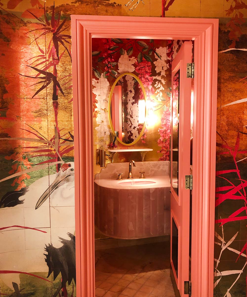 Looking through the doorway into the Ladies cubicle. Gold wallpaper overlaid with hand-painted bamboo and wisteria in pink and white. A large Asian Crane peeks out of the doorway.