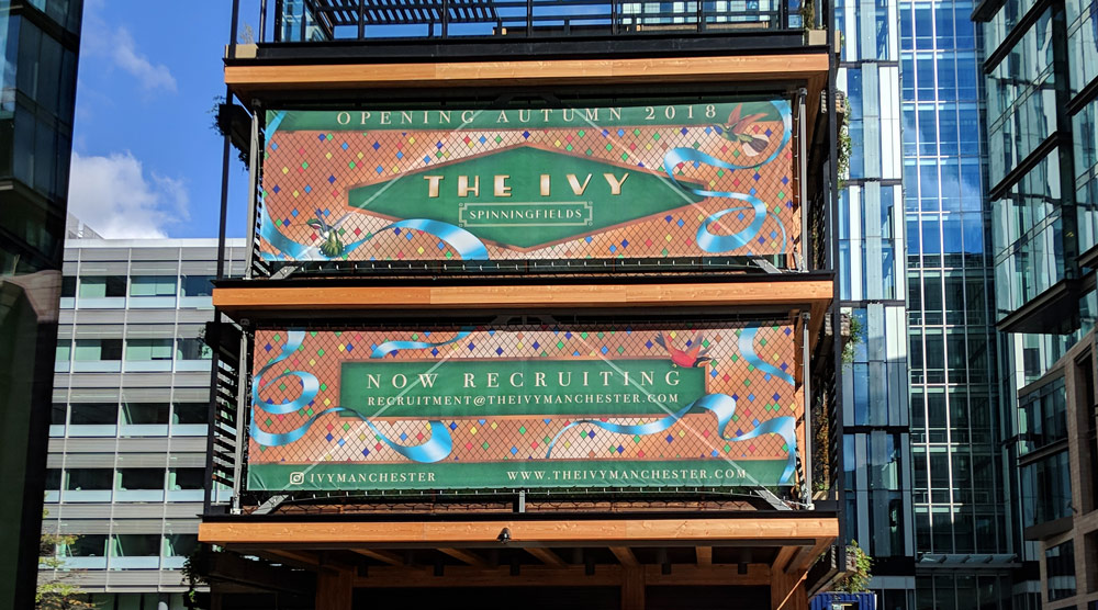 2 large format advertising banners displaying reservation and opening information stretched across the 2nd and 3rd floor windows of the building. The artwork displays the Ivy logo on a background of gold diamonds. Light blue ribbon and hummingbirds interact with the messaging.
