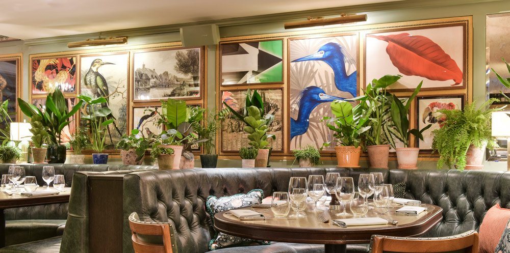 Restaurant panelling filled with contemporary artwork. Two Blue pelican heads are a large feature piece. Dining banquettes are in the foreground and many potted plants are positioned around the tables and in front of the framed artwork.