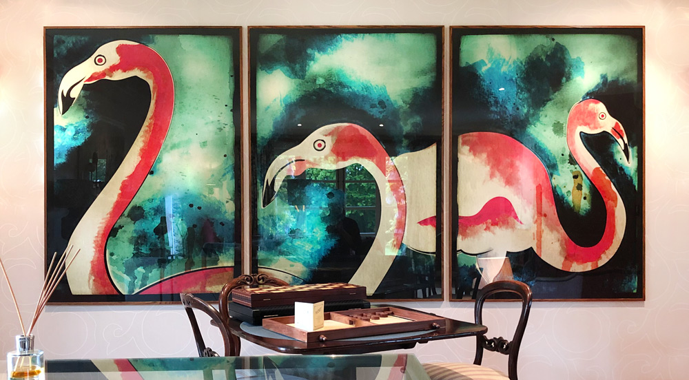 A set of three watercolour flamingoes against a teal and blue background. The artwork is split across a triptych layout and framed in a medium oak moulding.
