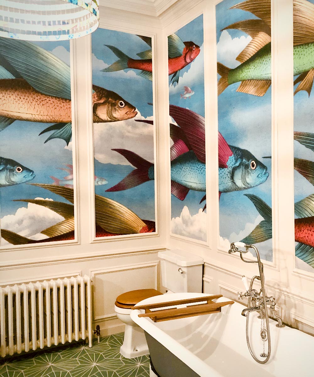 Bathroom panelling displays our flying fish wallpaper. Blue, red, orange and green fish with coloured wings fly across a cloudy blue sky. A bathtub and toilet are in the foreground and the floor is made of a green geometric pattern.