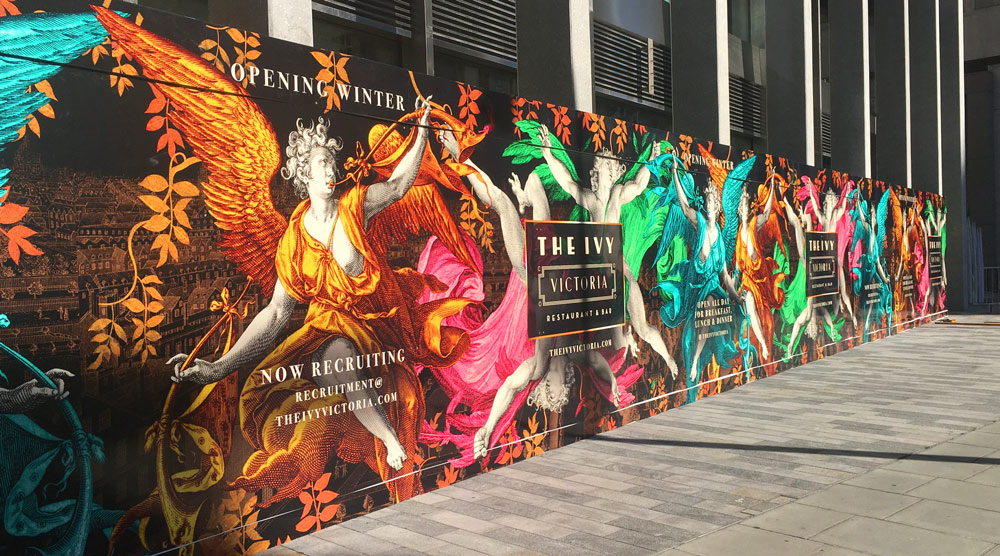 Hoarding artwork displays dramatic winged angels playing instruments over a nighttime view of London. The angel wings are coloured in vivid pinks, golds, cyan and green.